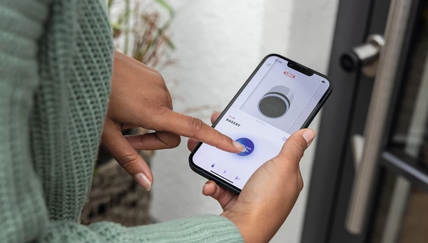 Woman holds smartphone with ABUS One app in her hand and opens the door in front of her with the CYLOX One © ABUS